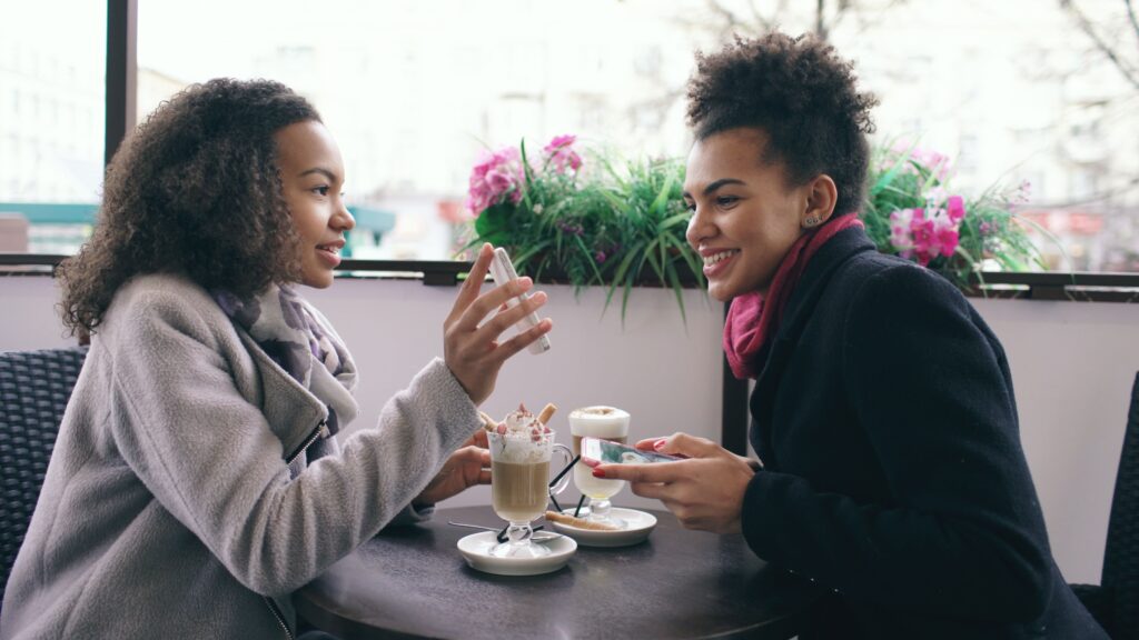Dos amigas sonríen mientras usan el celular juntas en una cafetería, reflejando el uso cotidiano y seguro de apps de pago entre personas conocidas